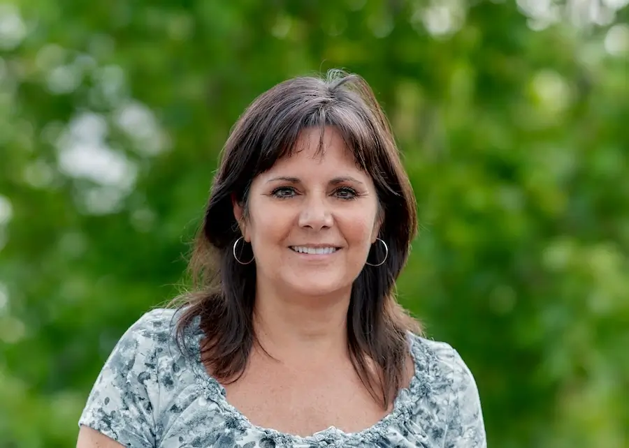 smiling woman with a gray shirt and earrings standing in front of trees